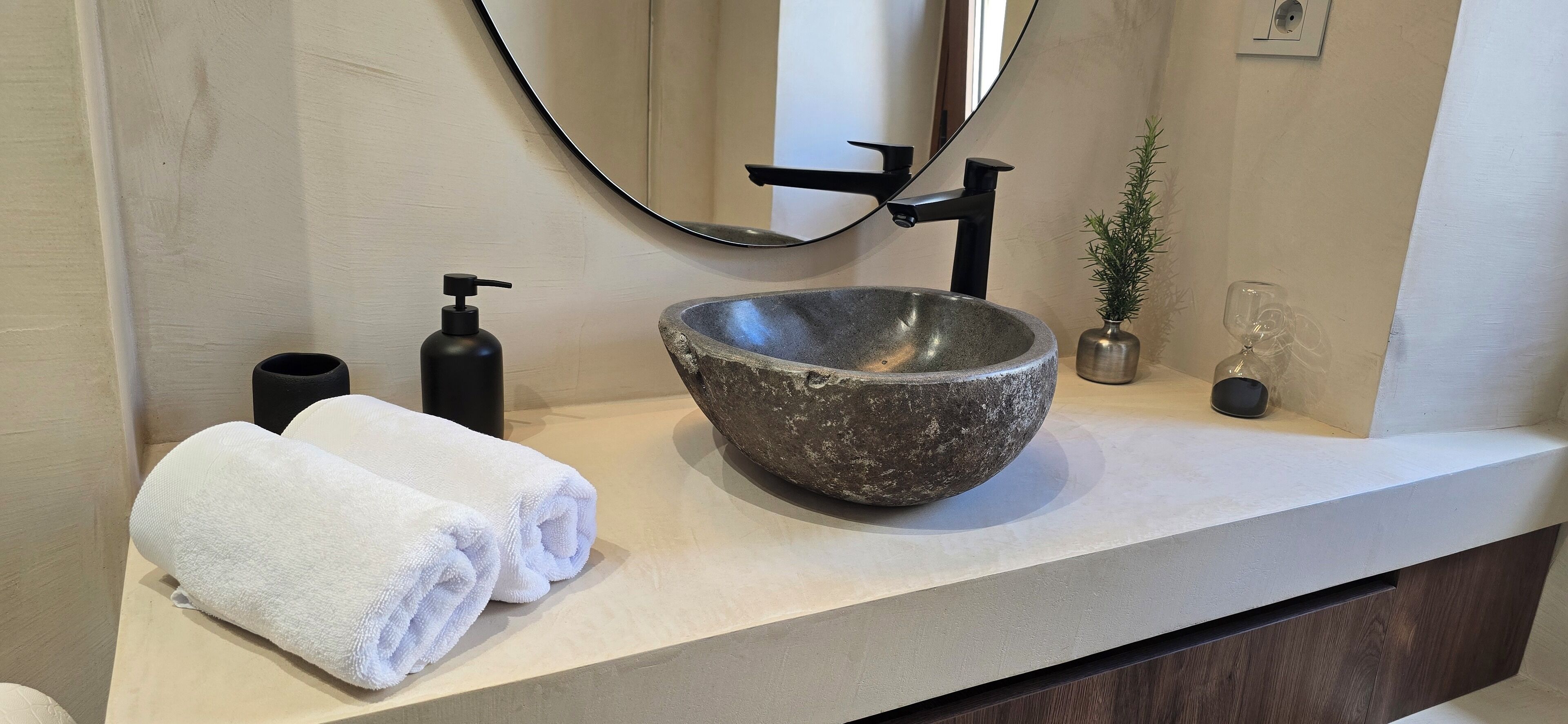 Une salle de bain avec un lavabo en pierre, un robinet noir, un miroir, une serviette, un distributeur de savon et une petite plante en pot.