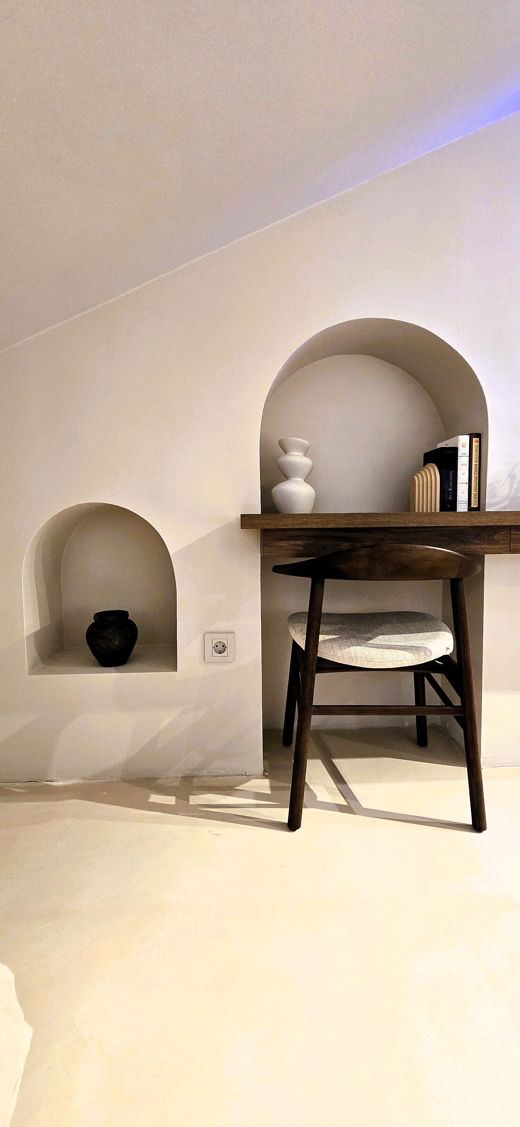 A minimalist room with a wooden table and a chair, a white vase, and books on a shelf.