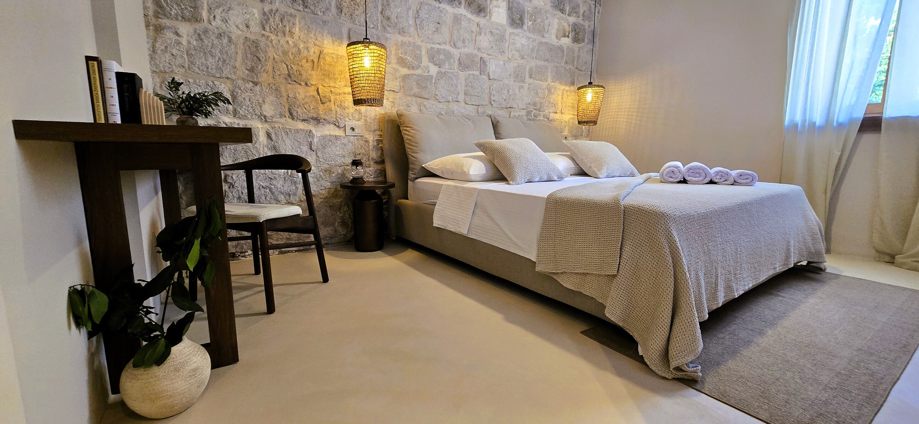 A bedroom with a stone wall, a bed with pillows and a blanket, a bedside table with a lamp, and a chair.