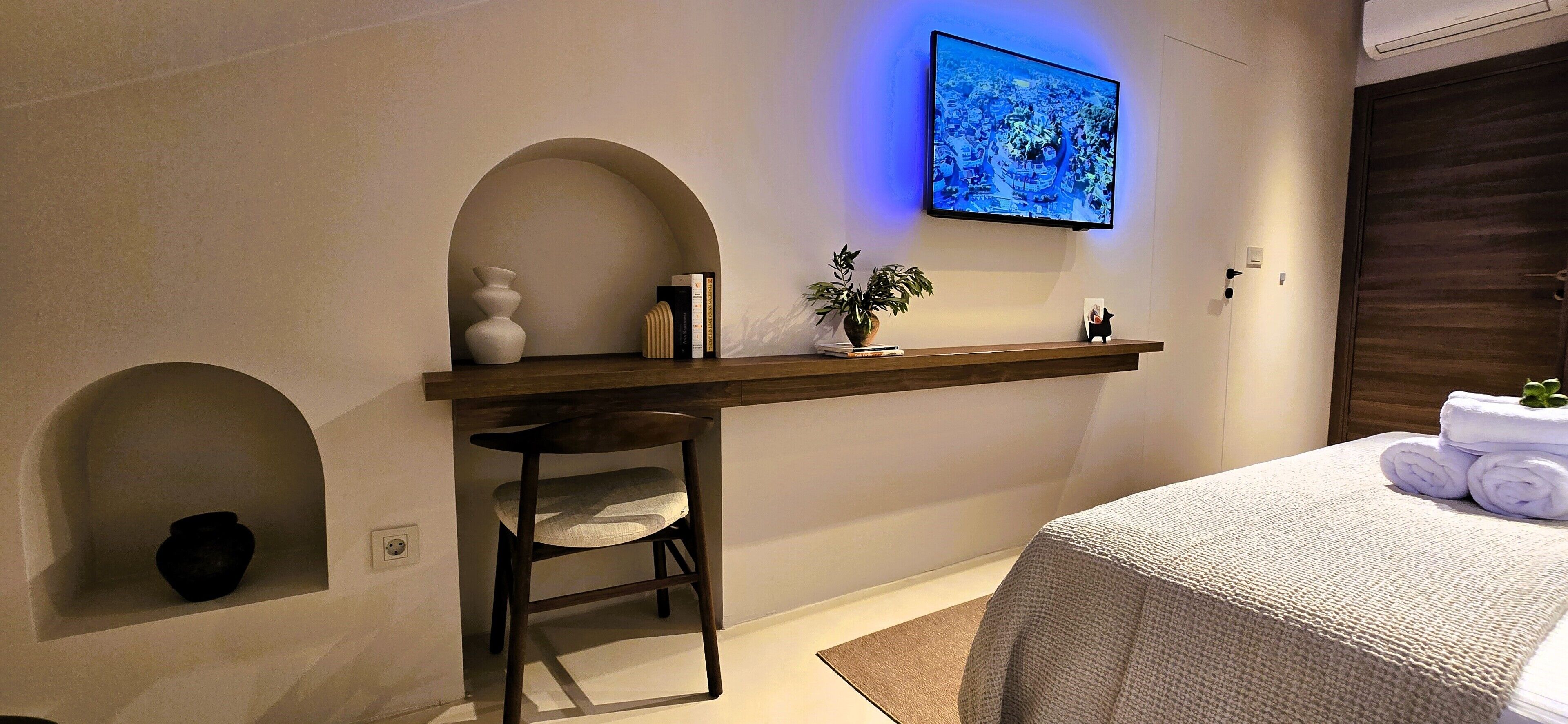 Ein Schlafzimmer mit einem Bett, einem an der Wand befestigten Fernseher, einem Regal mit Büchern und einer Pflanze sowie einem Sessel.