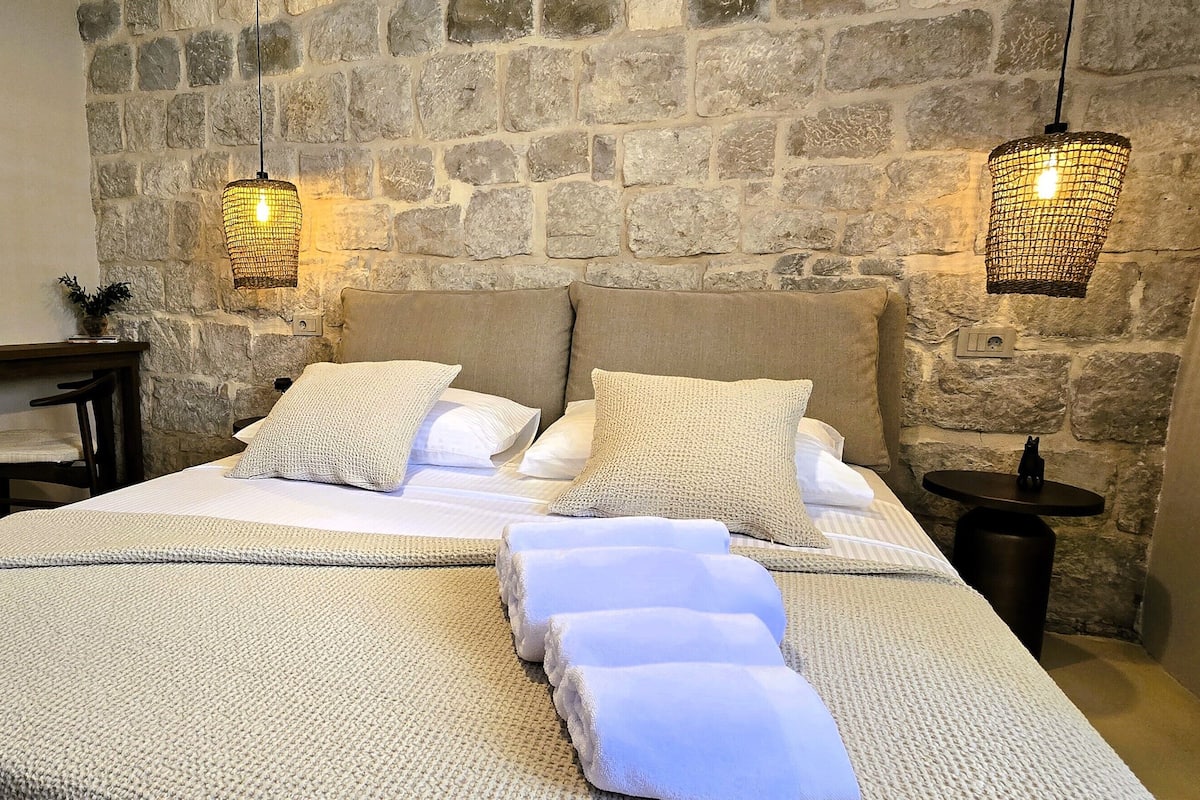 Una camera da letto con parete in pietra, un letto con lenzuola bianche, due lampade da comodino e un tavolino con una pianta.