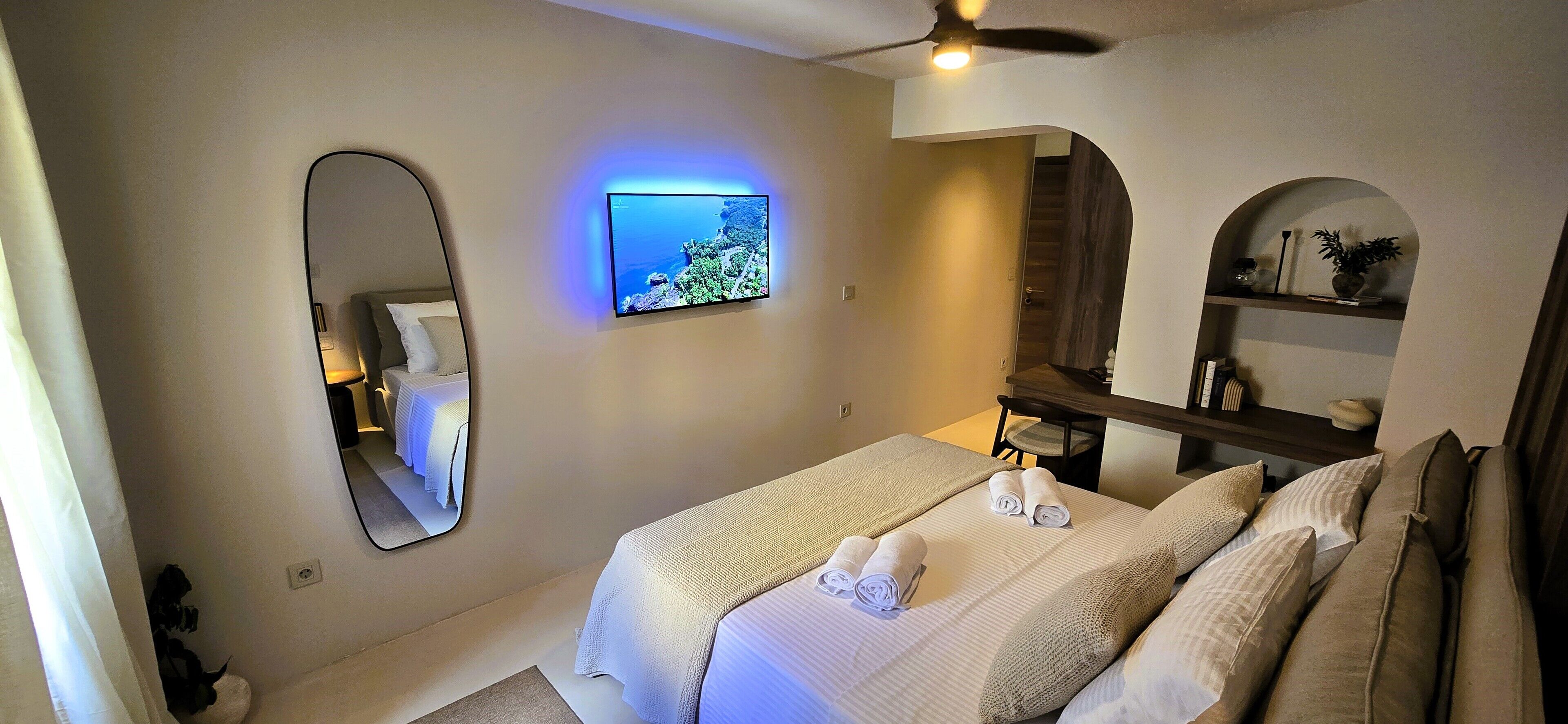 Habitación de hotel con cama, televisor montado en la pared, espejo, escritorio pequeño y estante con objetos decorativos.
