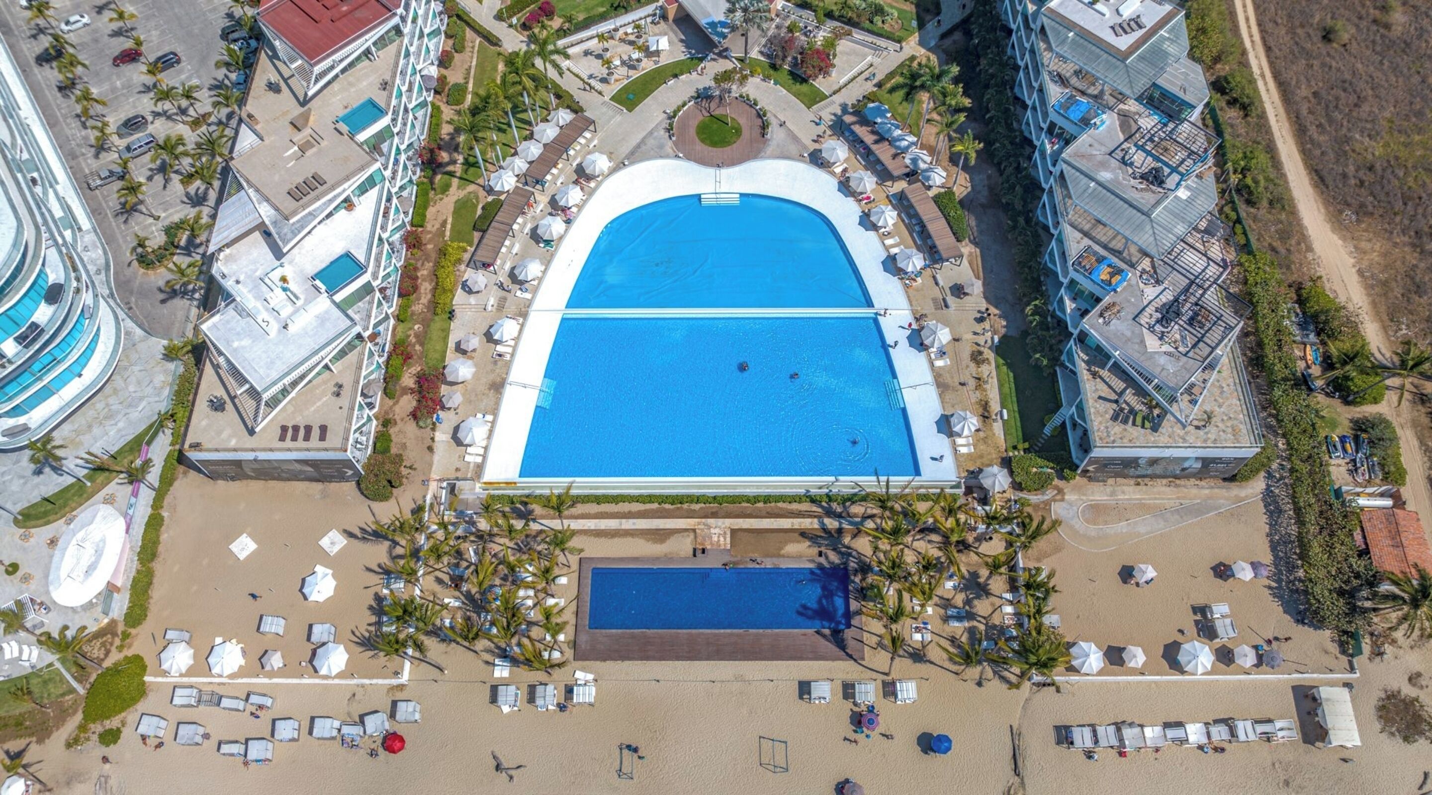 An aerial view of a resort with a central swimming pool, multiple beach areas with sun loungers, and surrounding buildings.