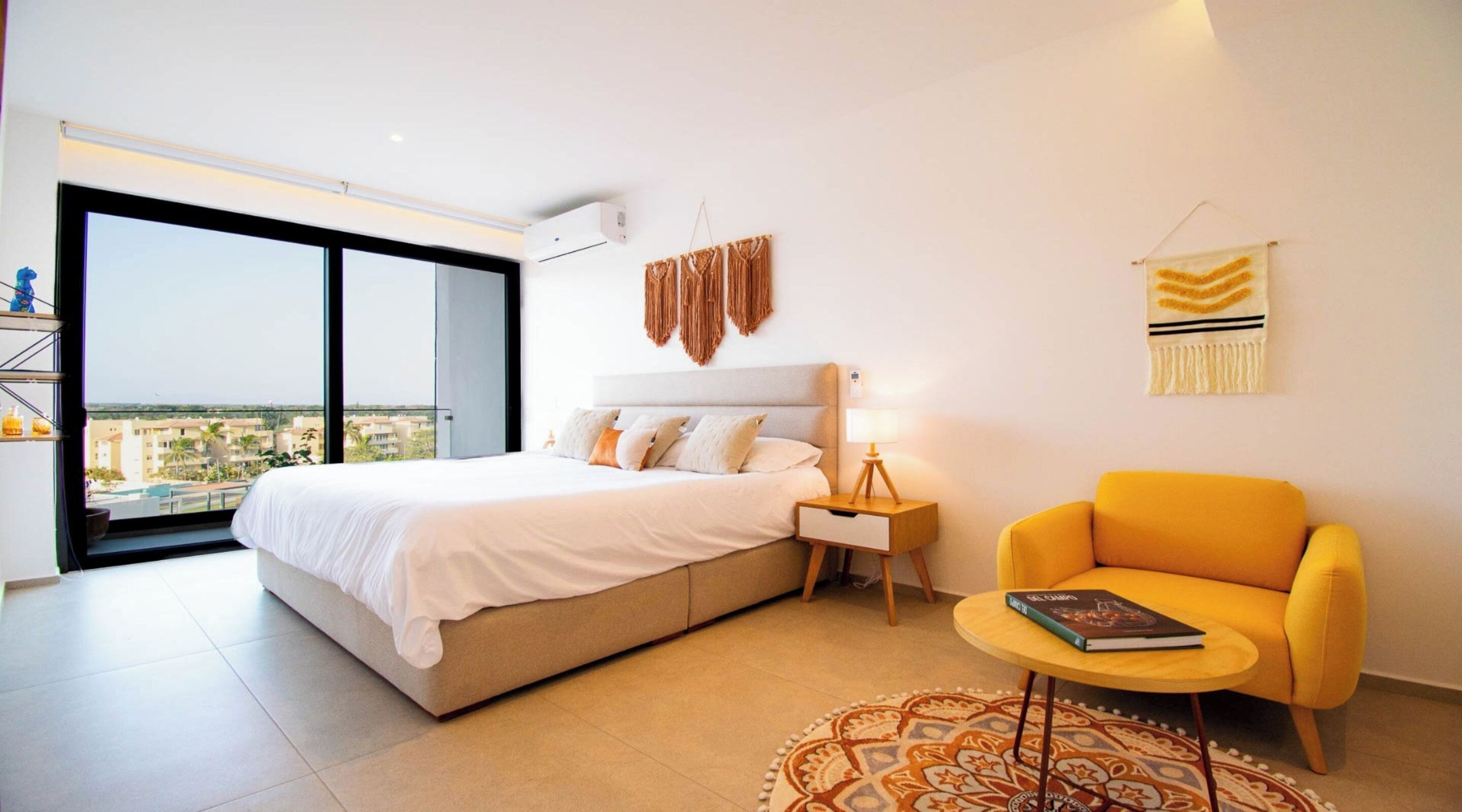A modern bedroom with a large bed, a yellow armchair, and a view of the city through a large window.