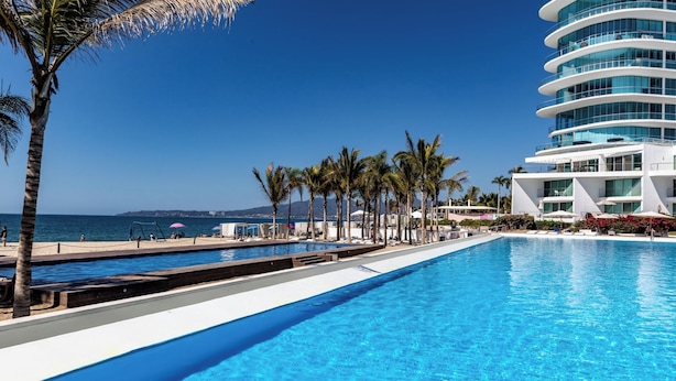 A pool area with a clear view of the ocean, palm trees, and a modern hotel building. - Condo Aria Ocean 614 by Kivoya (Nuevo Nayarit)