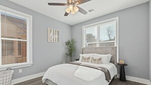 A bedroom with a large bed, a ceiling fan, a window with blinds, a small side table, and a plant. - Cozy 3BR Home - Fire Pit & Gated Parking in Midtown - Near Rhodes College & Zoo (Memphis)
