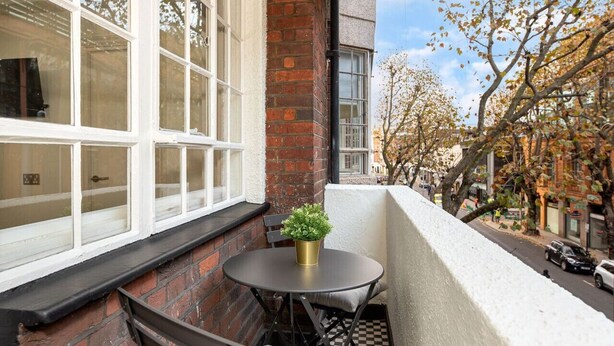 Un balcon avec une petite table et un pot de fleurs, donnant sur une rue bordée d’arbres aux couleurs automnales et de voitures garées.