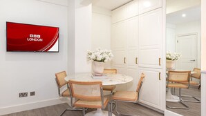 Un espace repas moderne avec une table ronde, quatre chaises en osier, un vase de fleurs et un téléviseur à écran plat diffusant la chaîne « BBC LONDON ».