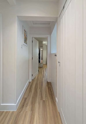 Um corredor com piso de madeira e paredes brancas, que dá para um quarto com uma porta.