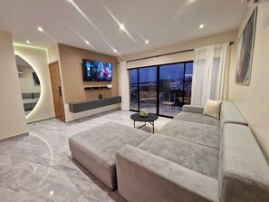 Een moderne woonkamer met een grote grijze modulaire bank, een flatscreen televisie bevestigd aan een houten paneel, en een groot raam met uitzicht op de stad.