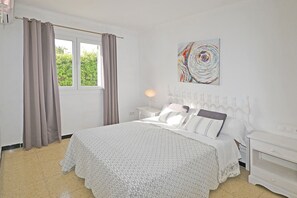 Ein Schlafzimmer mit einem Bett, weißen Nachttischen, einer Lampe und einem Fenster mit Vorhängen.