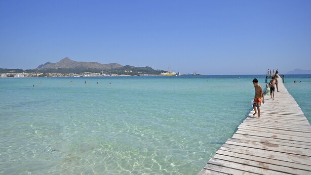 A wooden pier extending into clear turquoise water with a mountain in the background and a cityscape across the water.