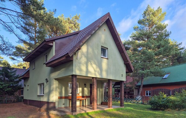 A two-story house with a green exterior and a brown roof, featuring a porch with a wooden bench and a small table.