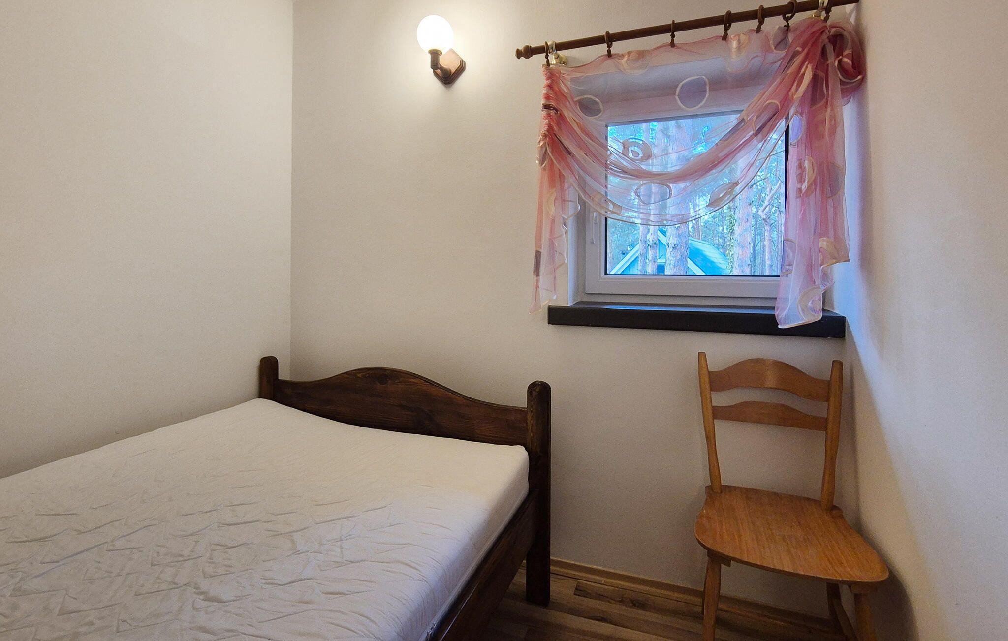 Une chambre à coucher avec un lit en bois, une chaise, une fenêtre avec des rideaux légers et un luminaire fixé au mur.
