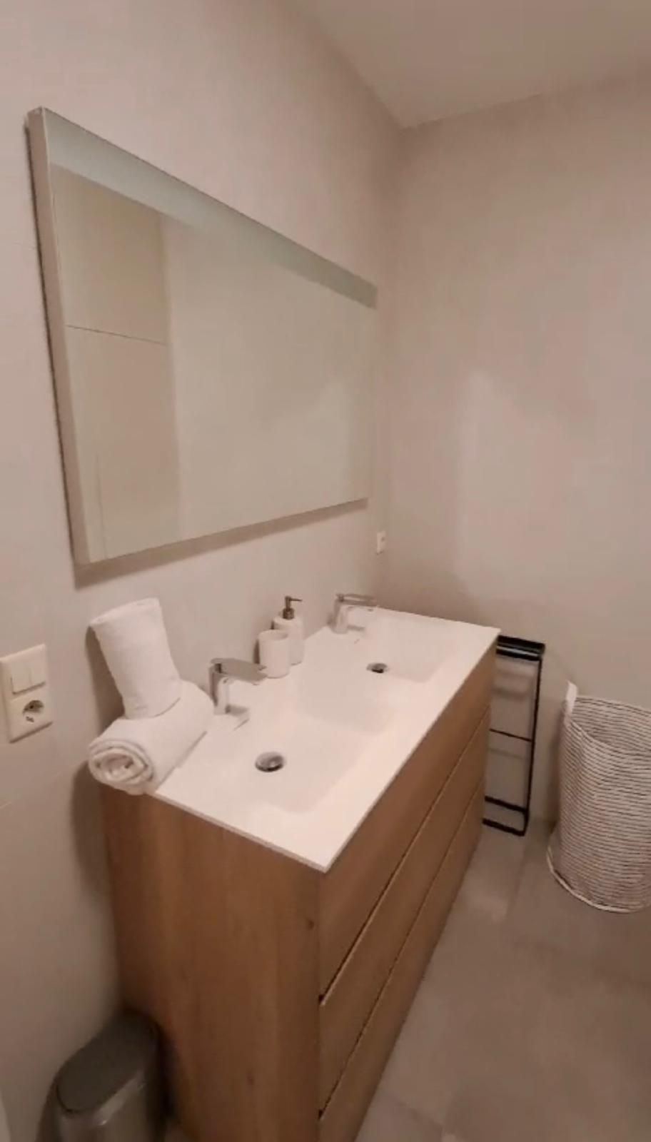 Une salle de bain avec un meuble-lavabo en bois, deux vasques, un miroir et une poubelle.