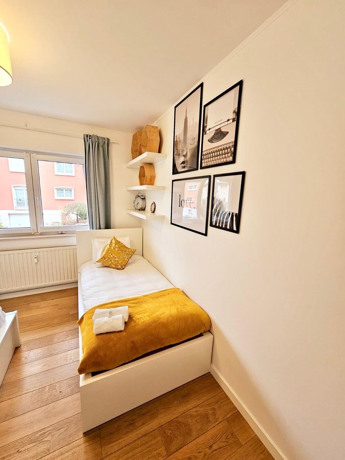 A neatly arranged bedroom with a bed, a bedside table, a window with curtains, and wall-mounted shelves with decorative items.