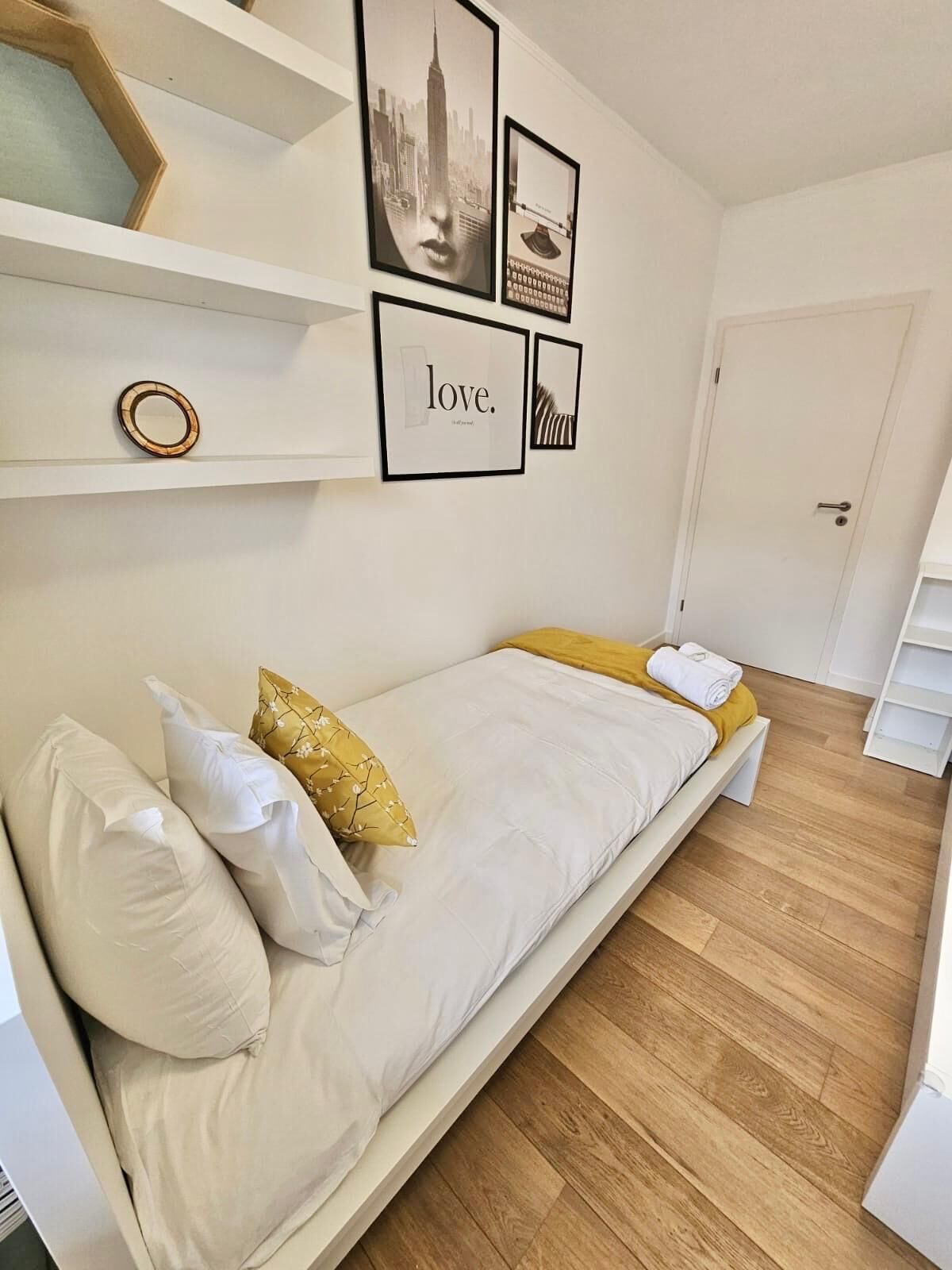 A bedroom with a bed, pillows, a shelf with framed pictures, and a door.