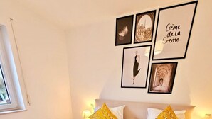 Una cama bien hecha con una manta amarilla y dos almohadas, una ventana con persianas y cuadros en la pared.