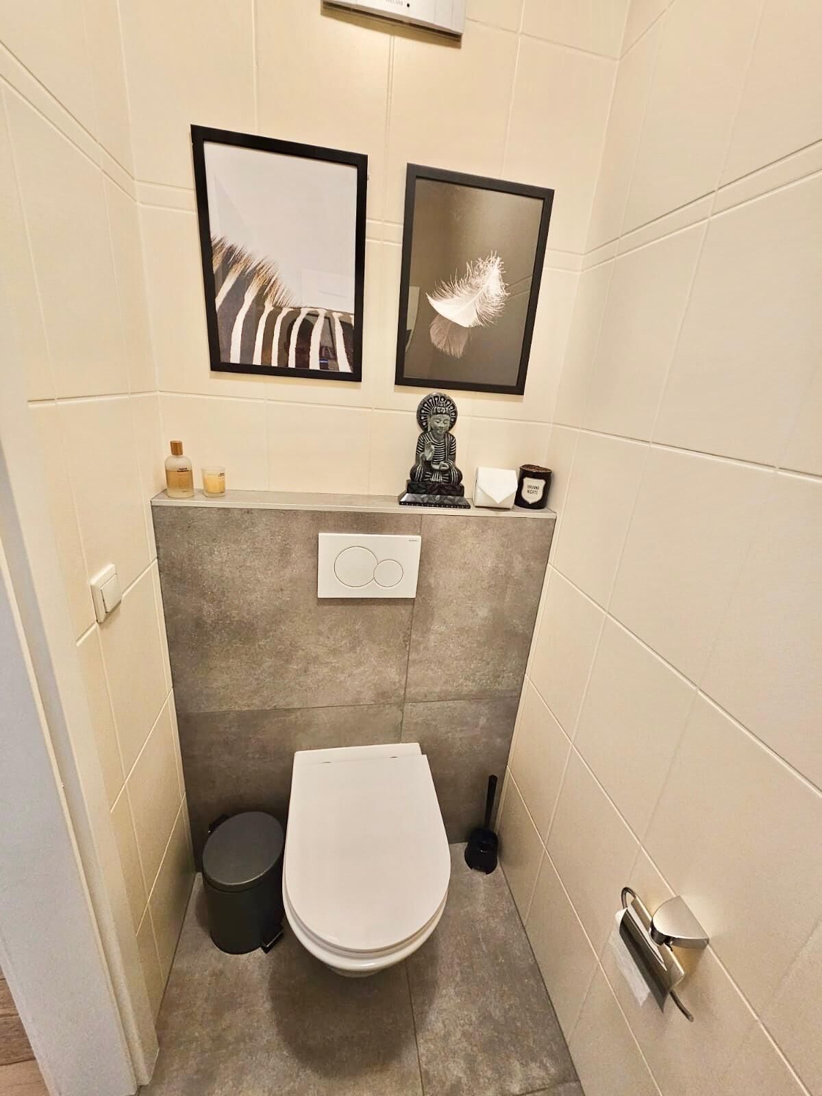 Et lille badeværelse med toilet, en lille skraldespand og en toiletpapirholder på væggen. Der er to billeder i rammer over toilettet.