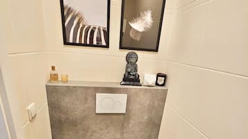 Et lille badeværelse med toilet, en lille skraldespand og en toiletpapirholder på væggen. Der er to billeder i rammer over toilettet.