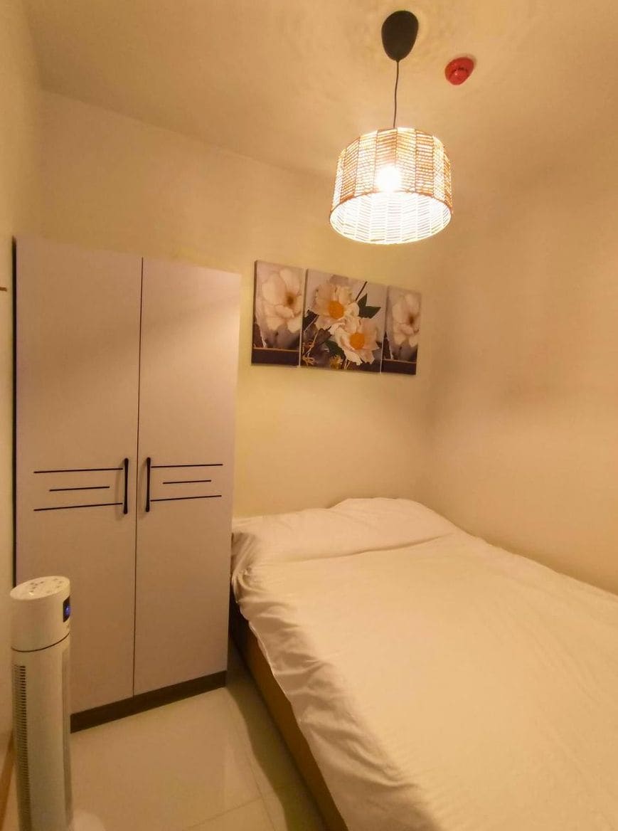 A small, single-bed room with a wardrobe, a wall-mounted light fixture, and a small air purifier.