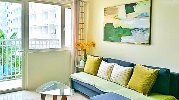 Un salon avec un canapé bleu, une table basse en verre, un ventilateur de plafond, un tableau et une vue sur un immeuble par la fenêtre.