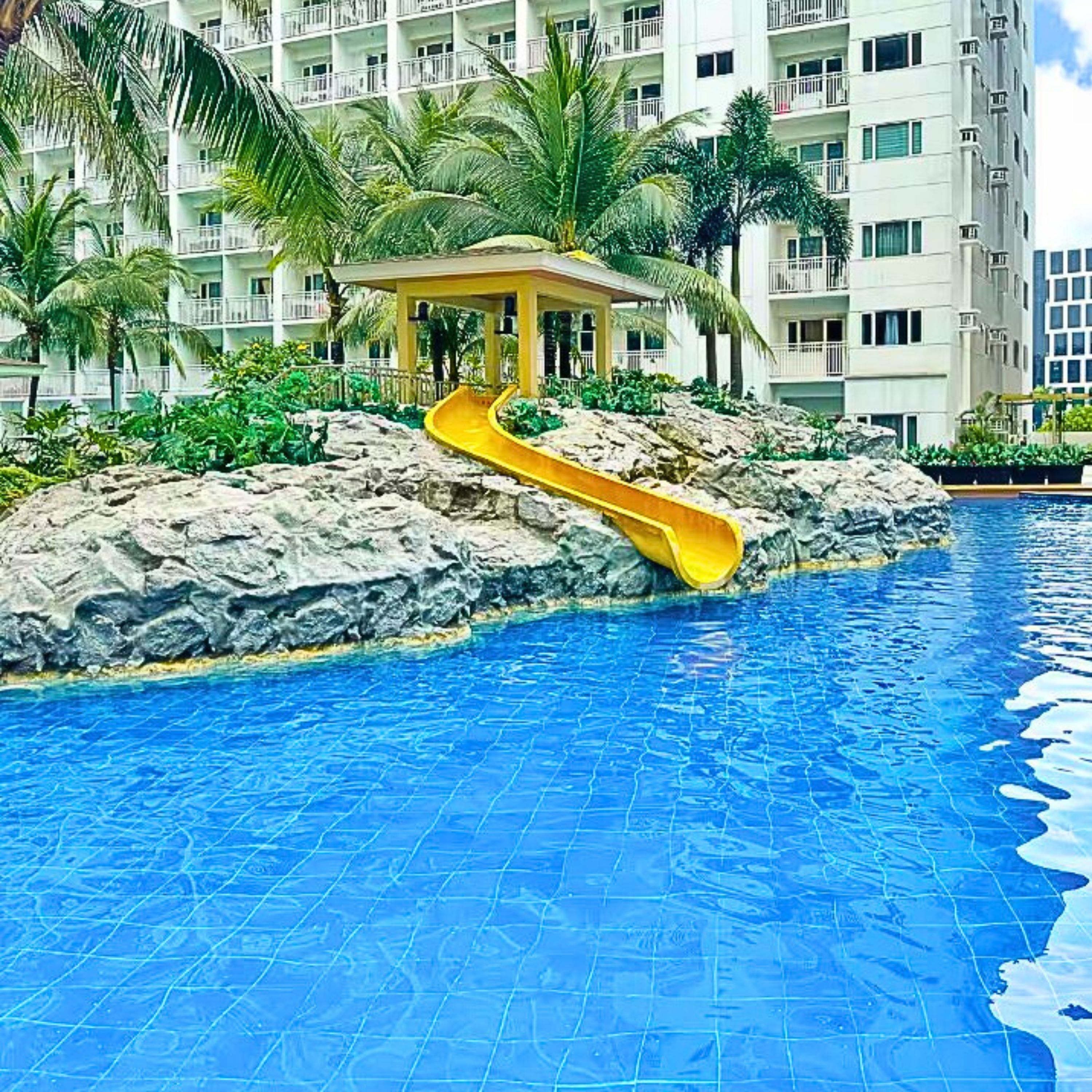A swimming pool with a water slide, surrounded by palm trees and a building with balconies.