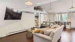 A farmhouse-style living room with a large TV, a sofa, a coffee table, and a kitchen area with a sliding door. - Charming 3bd Retreat– near Estes (Drake)