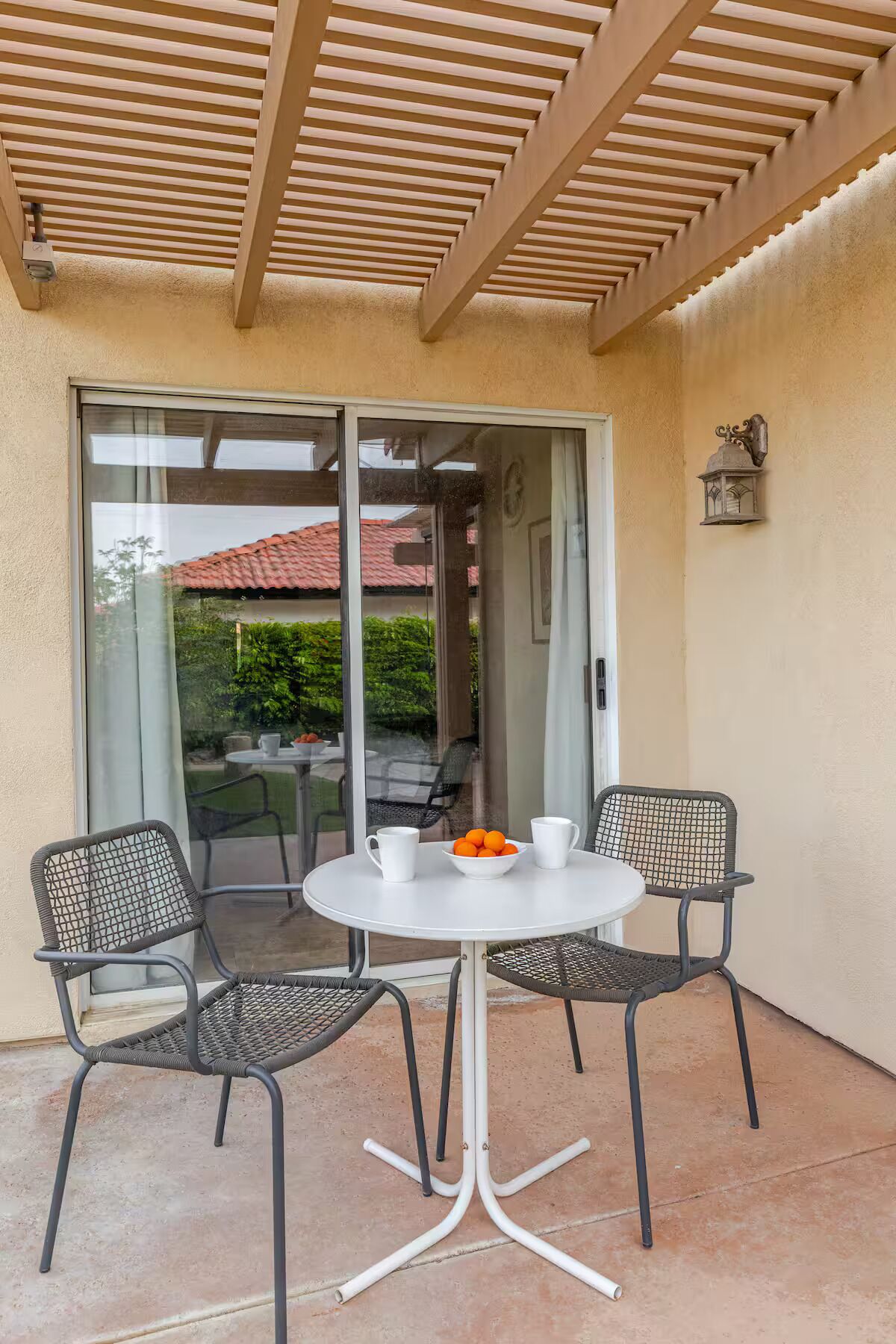 An outdoor patio with a round table and four chairs, a glass door leading to a garden, and a wall-mounted lantern.