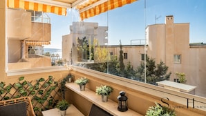 A balcony with potted plants, a wicker chair, and a view of buildings and the sea.