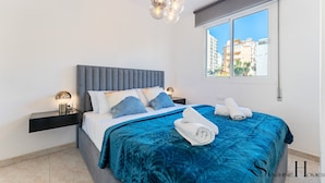 A modern bedroom with a grey upholstered headboard, a blue patterned bedspread, and a view of buildings through a window.
