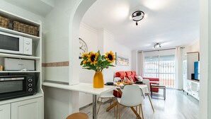 A modern kitchen with white cabinets, a microwave, and a toaster. There is a dining area with a white table, red chairs, and a vase of sunflowers. - 50 Meters from the Beach, 1 Bedroom with Parking REF013 (Torremolinos)
