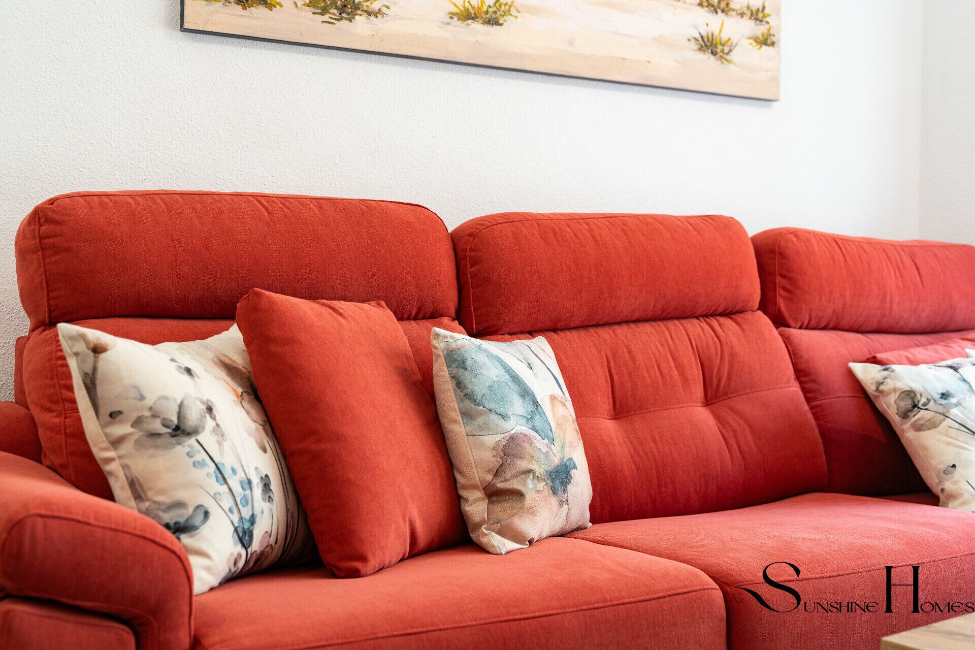 A red sofa with floral cushions and a painting above it.