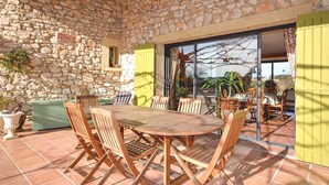Un patio con paredes de piedra, una mesa y sillas de madera, un banco verde y un ventanal de vidrio que da a un espacio exterior con plantas y vistas.