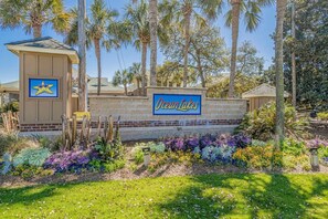 A sign for Ocean Lakes with a star logo, surrounded by palm trees and a landscaped garden. - Ocean Lakes Cottage, Covered Patio, Grill and Beach! (Myrtle Beach)