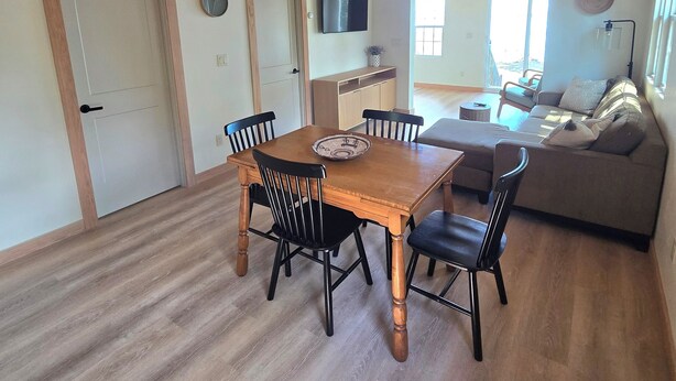 A dining area with a wooden table and chairs, a television on the wall, and a view of the living room with a sofa and a window. - The Lily Pad: Newly Renovated, Lakefront Retreat on Shawano Lake, Sleeps 6 (Shawano)