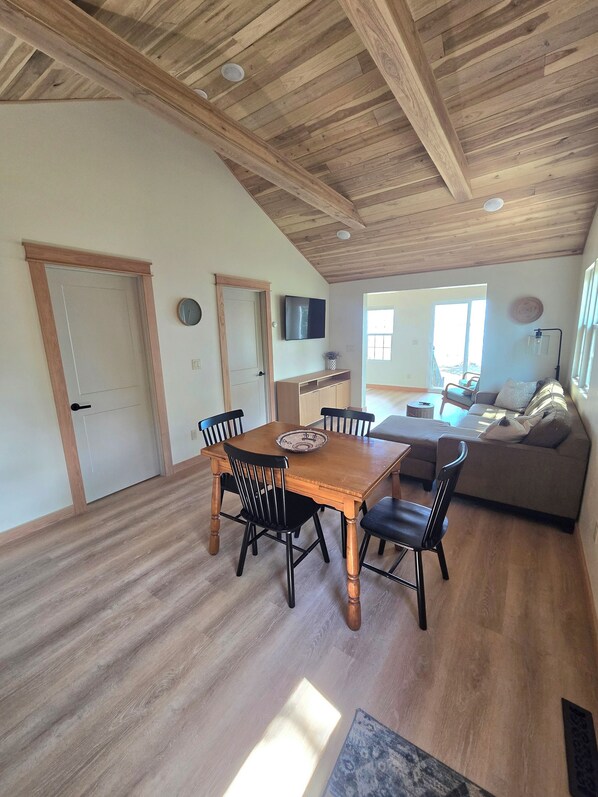 En matplats med ett träbord och stolar, en tv på väggen och utsikt över vardagsrummet med en soffa och ett fönster.