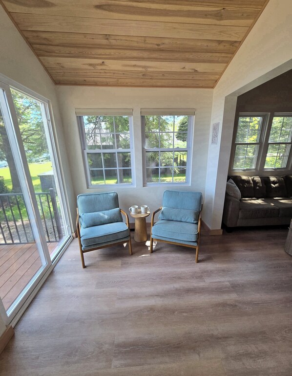 Una habitación con techo de madera, ventanas amplias, piso de madera, dos sillas azules, una pequeña mesa redonda con tazas y un sofá marrón.