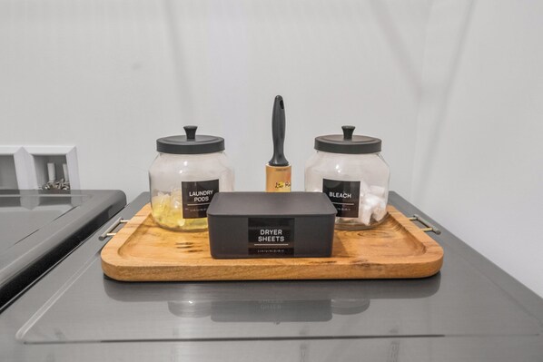 A kitchen counter with a wooden tray holding a black box labeled 'DRYER SHEETS' and two glass jars with lids, one labeled 'LAUNDRY PODS' and the other 'BLEACH'.