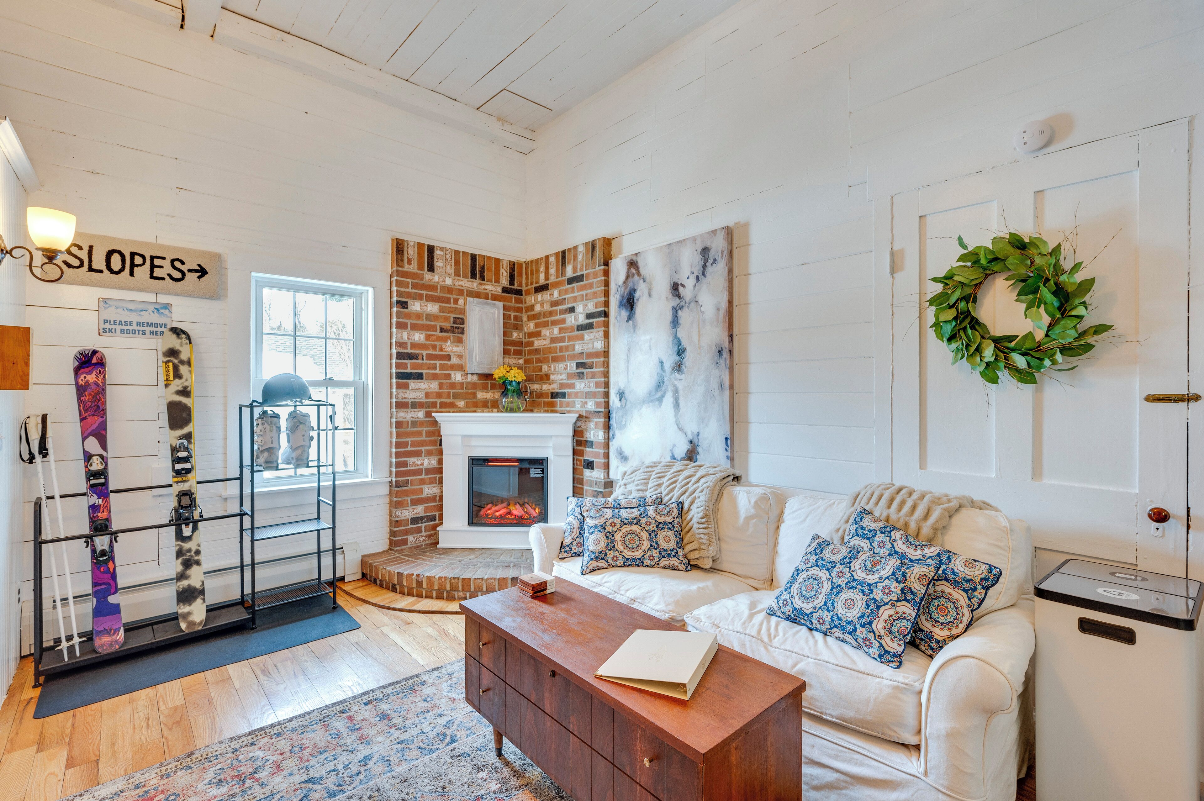 Un salon chaleureux avec une cheminée, un canapé blanc agrémenté de coussins à motifs, une table basse et un porte-chaussures de ski.