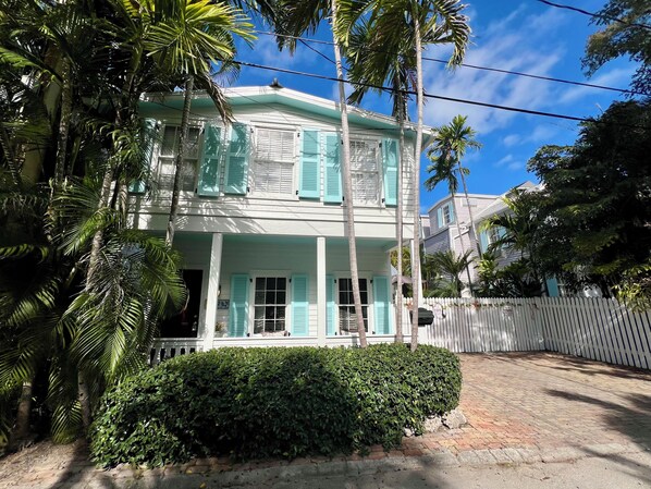 A two-story house with a white exterior and turquoise shutters, surrounded by lush greenery and a white picket fence. - Seaport Serenity Cottage - Quiet and Quaint (Key West)