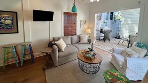 A living room with a grey sofa, a coffee table, a TV mounted on the wall, a ceiling fan, and a view of the backyard through a sliding door. - Seaport Serenity Cottage - Quiet and Quaint (Key West)