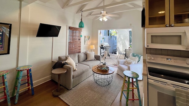 A living room with a grey sofa, a round coffee table, a TV mounted on the wall, and a ceiling fan. - Seaport Serenity Cottage - Quiet and Quaint (Key West)