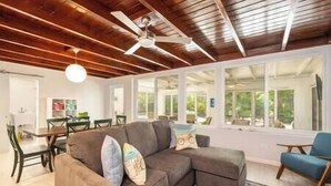 A spacious living room with a large sectional sofa, a ceiling fan, and large windows offering a view of the outdoors. - Plumeria Paradise, Your Tropical Retreat Close to the Beach (Key West)