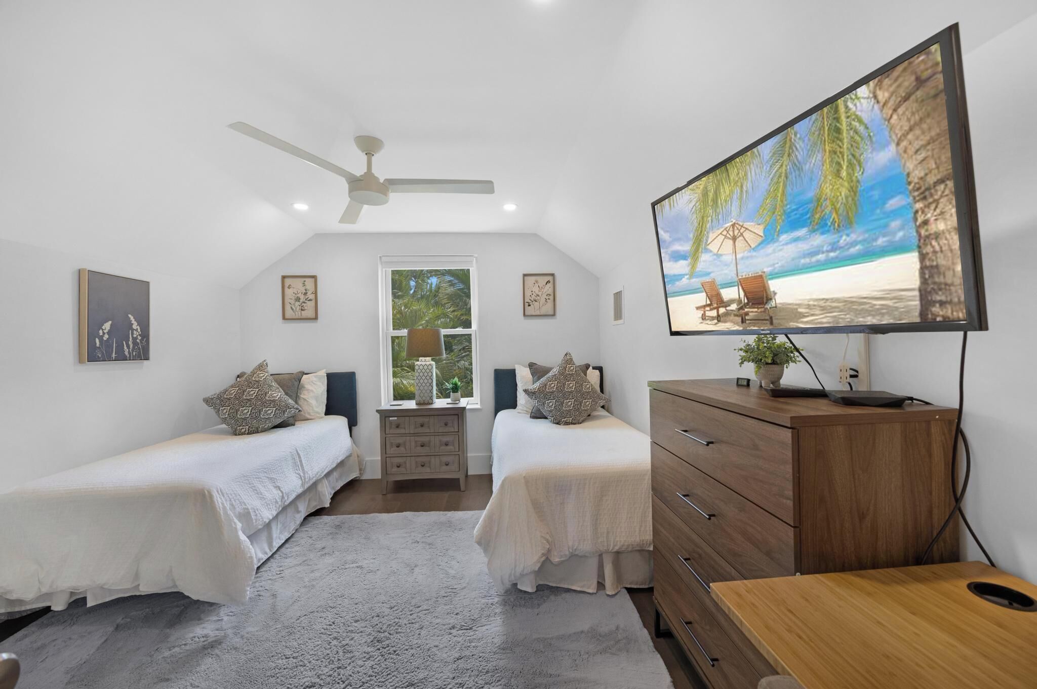 A bedroom with two single beds, a ceiling fan, a TV displaying a beach scene, a wooden dresser, and a window with a view of palm trees.