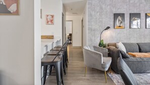 Een moderne woonkamer met een grijze bank, een grijze fauteuil en een houten eettafel met barstoelen. Er hangen abstracte kunstwerken en een poster aan de muur.