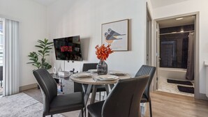 Een moderne eetruimte met een ronde tafel, zwarte stoelen, een vaas met oranje bloemen, een flatscreen televisie en een ingelijste foto aan de muur.