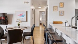 Eine moderne Küche mit weißer Arbeitsplatte, Spüle und einer hölzernen Bank. Es gibt einen Essbereich mit Tisch und Stühlen sowie ein gerahmtes Kunstwerk an der Wand.