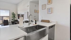 Eine moderne Küche mit Spüle aus Edelstahl, weißen Arbeitsplatten und einem Holzschnittbrett. An der Wand befinden sich gerahmte Kunstwerke und auf dem Balkon ein Blumentopf.