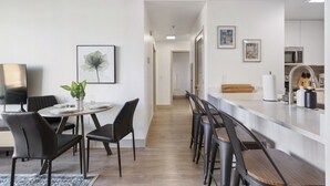 Moderni keittiö, jossa on valkoinen työtaso, ruostumattomasta teräksestä valmistetut kodinkoneet ja puinen ruokailutila, jossa on mustat tuolit.